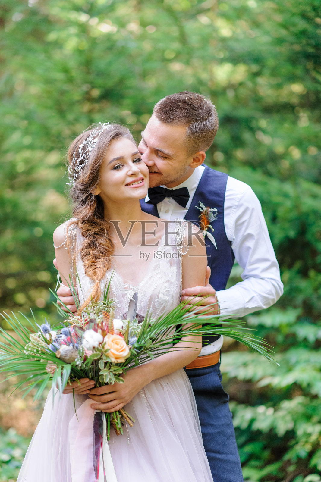 一对迷人的新婚夫妇，幸福快乐的时刻。一男一女穿着节日的衣服刮胡子亲吻。波希米亚风格的婚礼在森林里的新鲜空气中举行。照片摄影图片