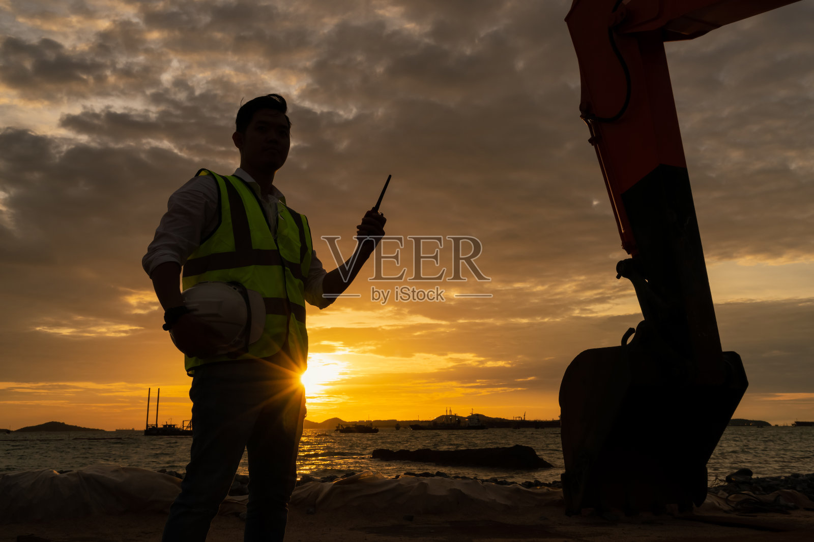 日落时分，建筑工程师在海边的建筑工地与挖土机保持无线电通讯。工程师的概念。照片摄影图片