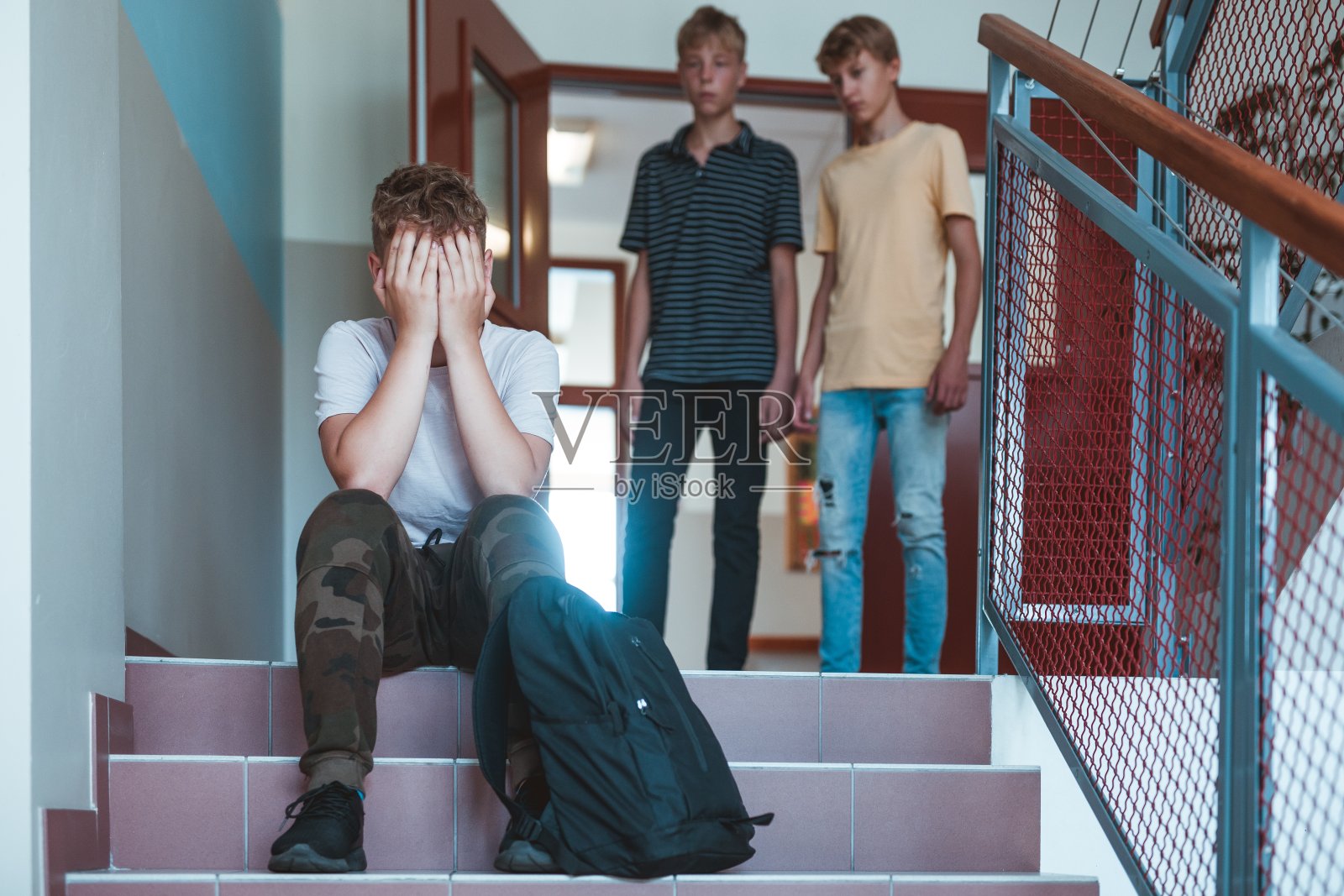 悲伤的孩子背着背包坐在学校的楼梯上，两个同学在他后面照片摄影图片