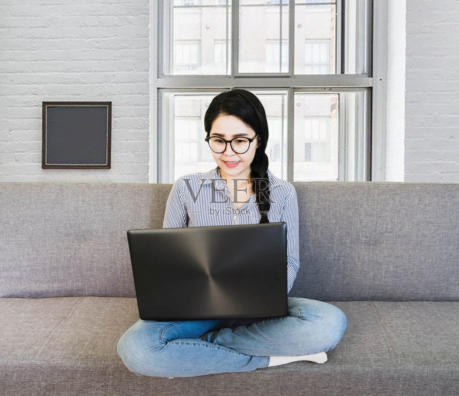 选择性聚焦年轻美丽的亚洲女子穿着蓝色条纹衬衫和眼镜坐在窗前的灰色沙发上，以网上工作的生活方式在家里用笔记本电脑放松照片摄影图片
