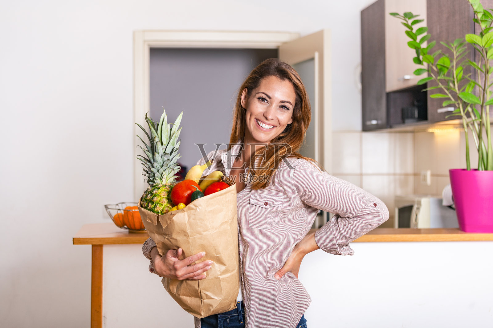 带一个健康的袋子。裁剪的图像美丽的年轻女子在围裙拿着纸购物袋充满新鲜蔬菜和微笑，而站在厨房前照片摄影图片