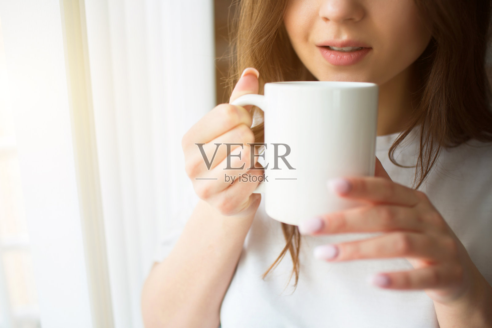 隔离期间有个年轻女人在厨房站在有白色窗帘的窗户前，拿一个盛有茶或咖啡的杯子。一个人喝闷酒。平静安宁的女人。减少视图。照片摄影图片
