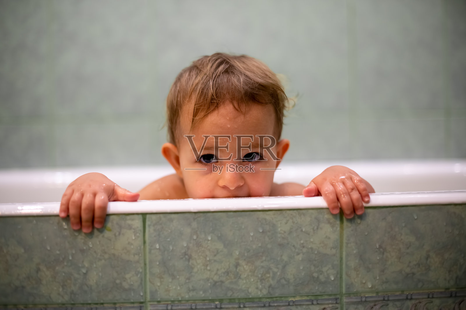 可爱的白人婴儿从浴缸里探出头来，把手放在浴缸边，看着镜头咬它。背景是一个模糊的绿色浴室。特写,软焦点照片摄影图片