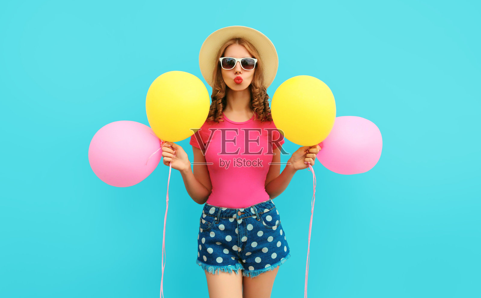 夏天五颜六色的愉快的形象可爱的年轻女子与黄粉色的气球有乐趣穿着短裤和草帽在蓝色的墙壁背景照片摄影图片
