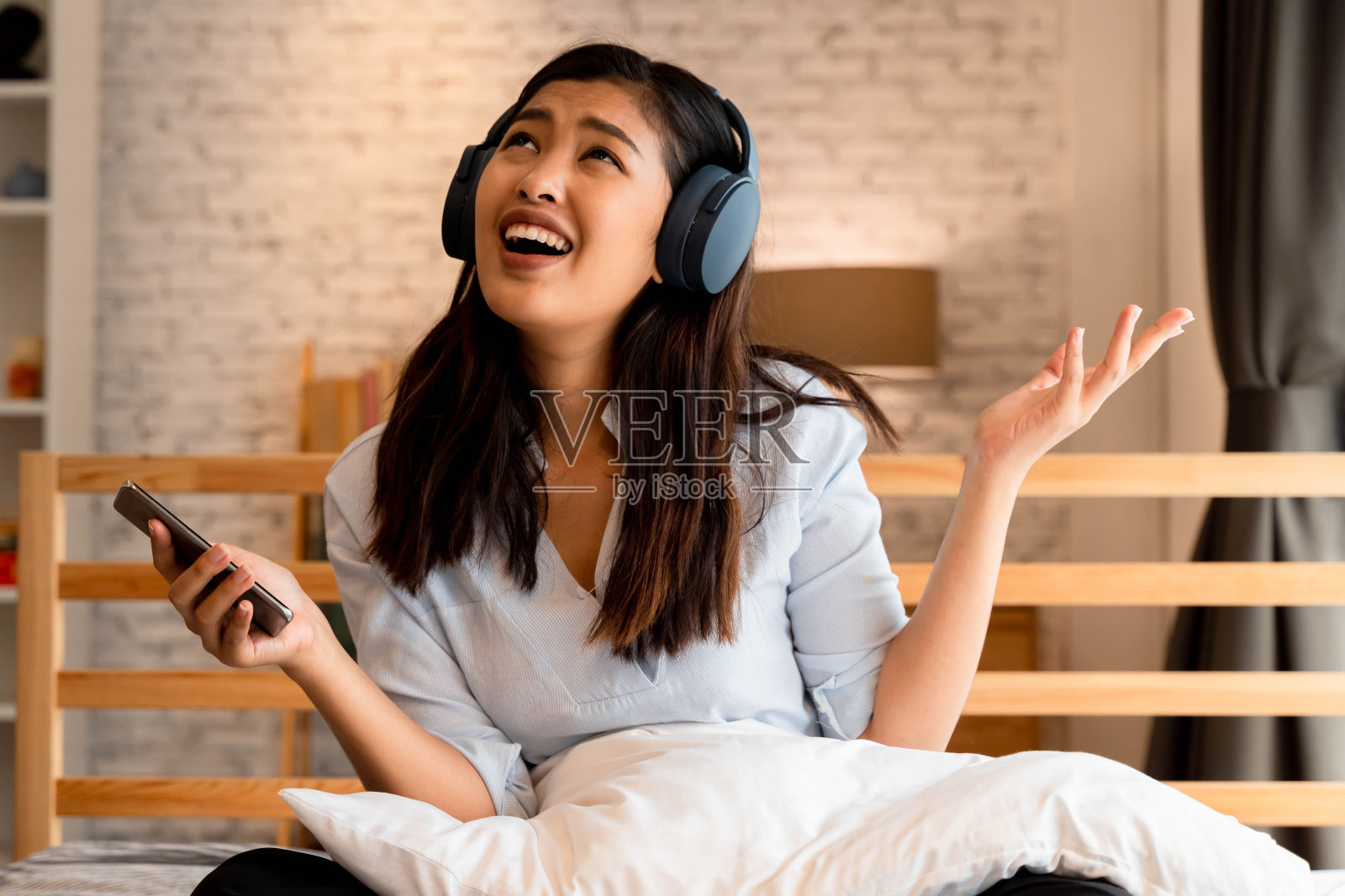 快乐的年轻亚洲女人戴着耳机唱歌和听音乐，而坐在床上，在家里拿着手机照片摄影图片