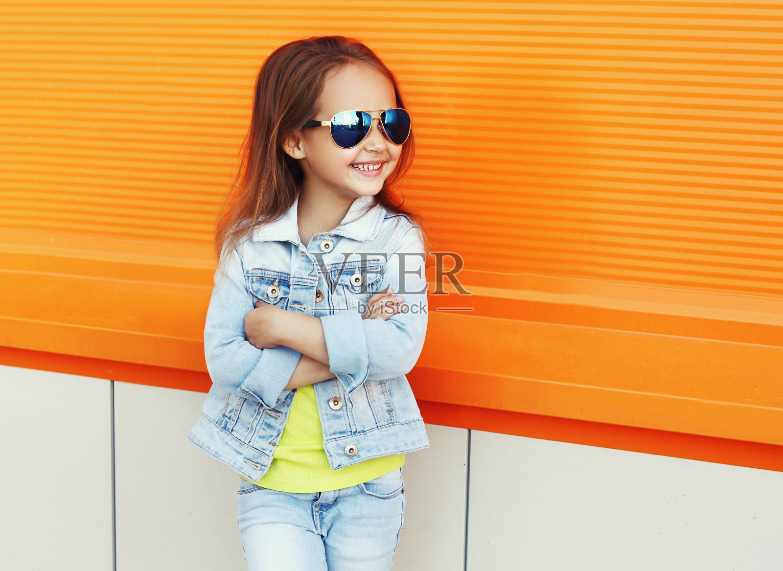一个小女孩的肖像，戴着太阳镜，牛仔裤夹克在城市街道上橙色的背景照片摄影图片