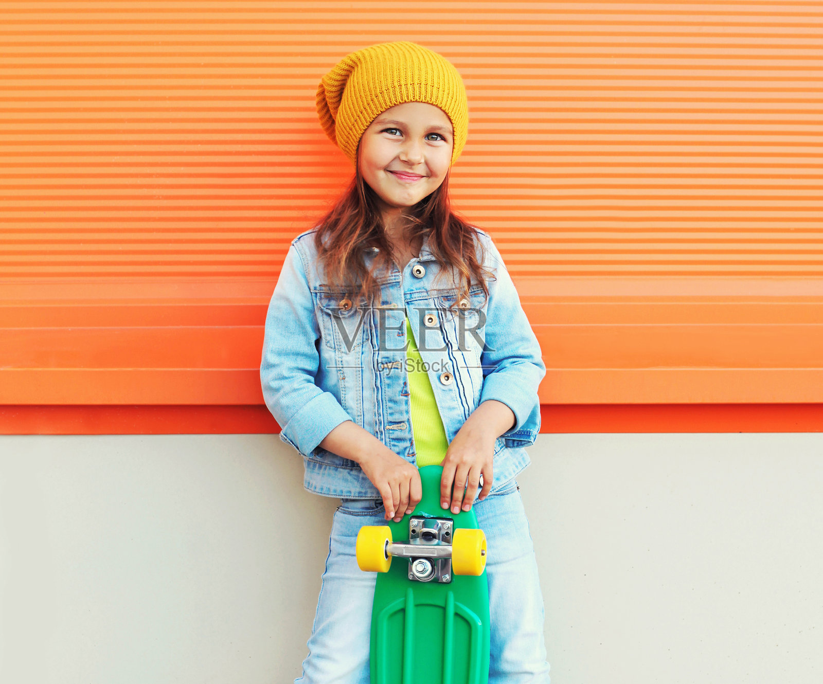 时尚微笑的小女孩的肖像与滑板在城市街道上的橙色墙背景照片摄影图片