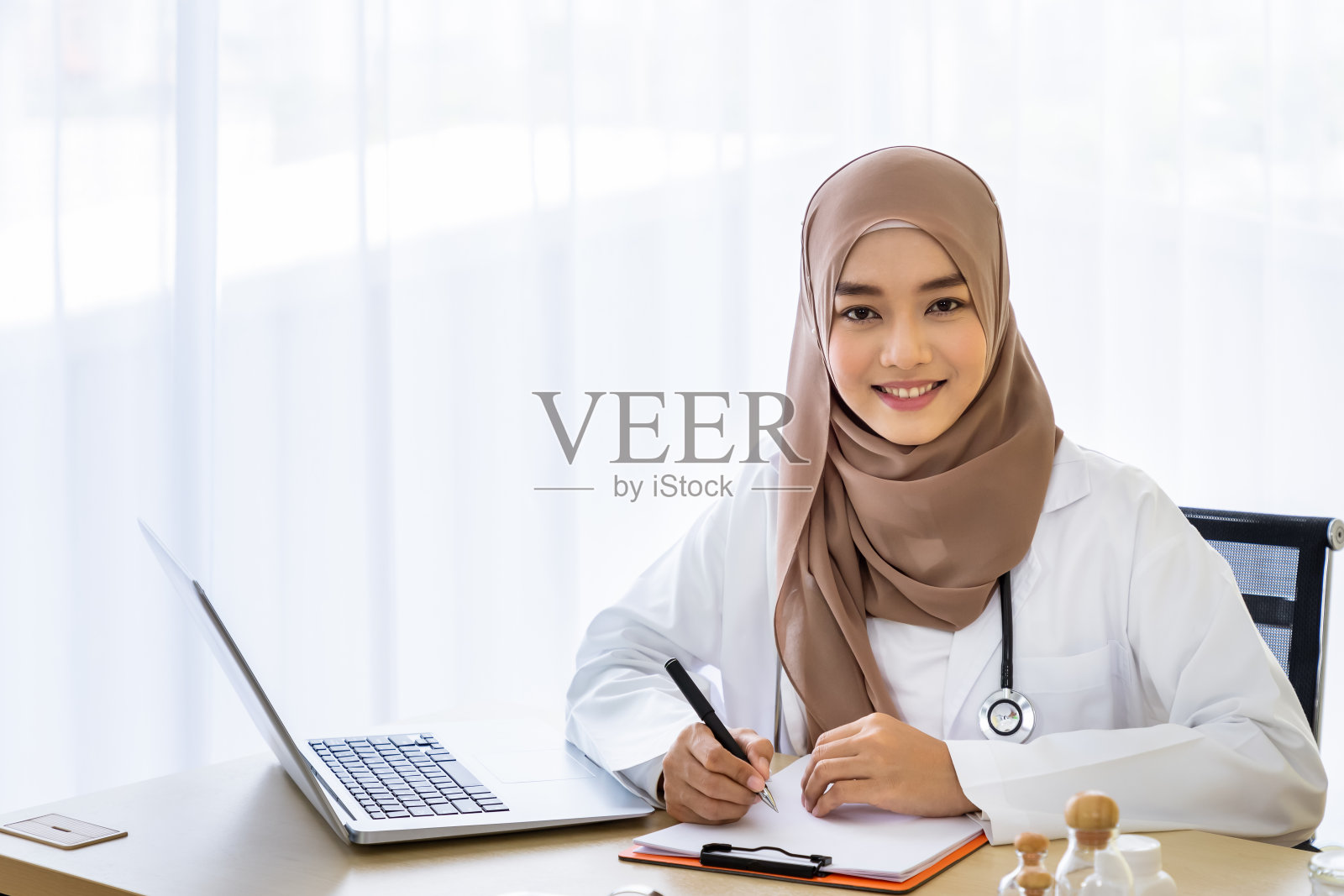 自信的穆斯林女医生坐在办公桌前，用笔记本电脑在笔记本上写在诊所医院办公室;看相机照片摄影图片