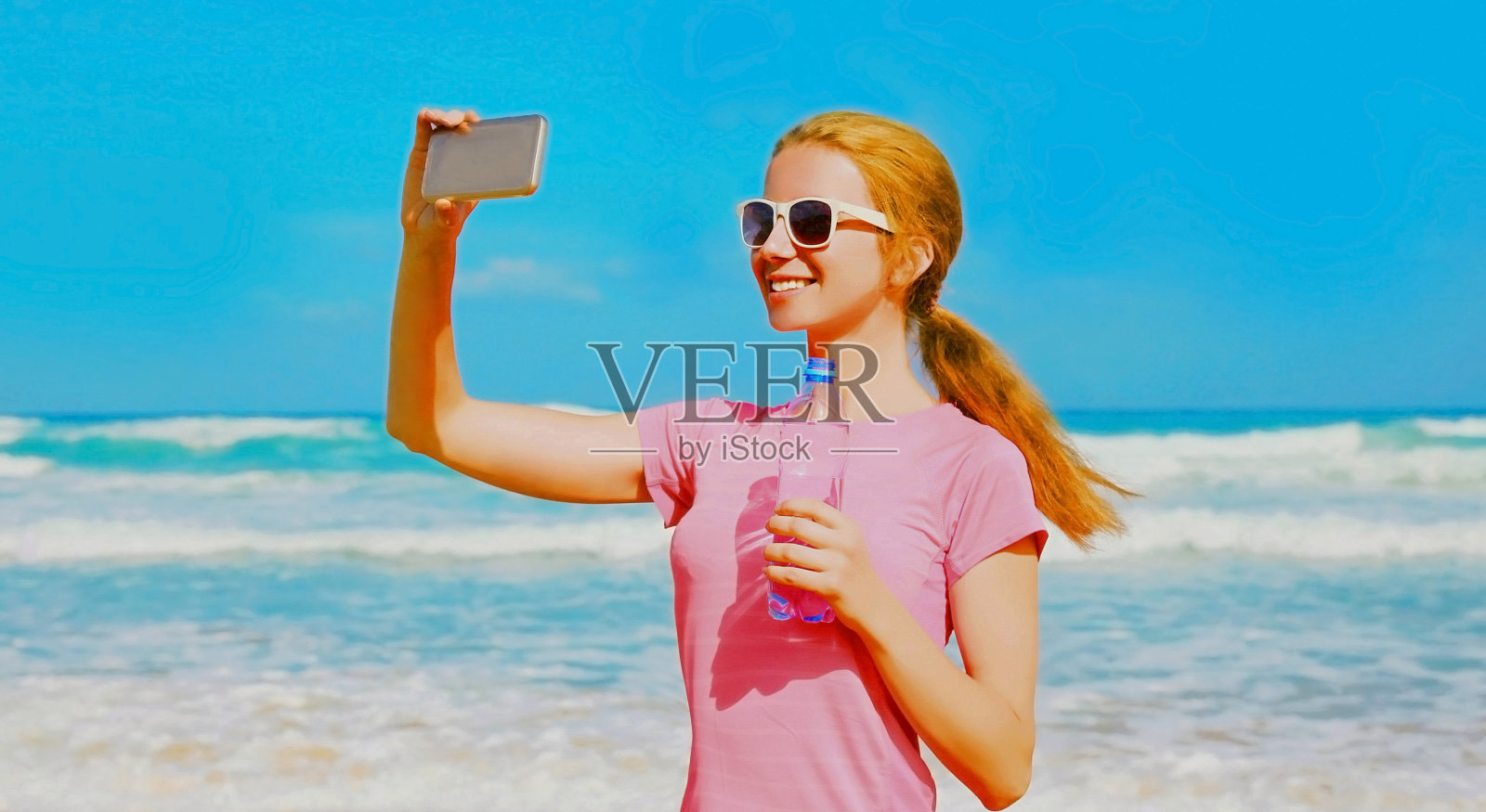 健身快乐微笑的女人拿着一瓶水在海滩上用智能手机自拍照片摄影图片