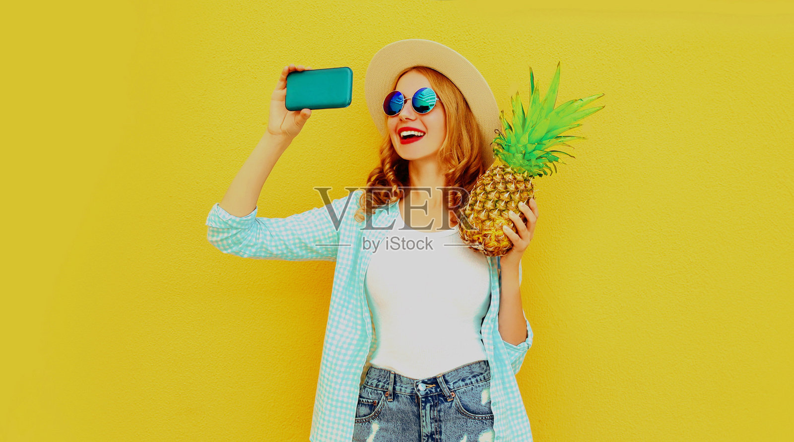 一个快乐的微笑女人与菠萝的夏季肖像用智能手机自拍照片戴着草帽，太阳眼镜在多彩的黄色背景照片摄影图片