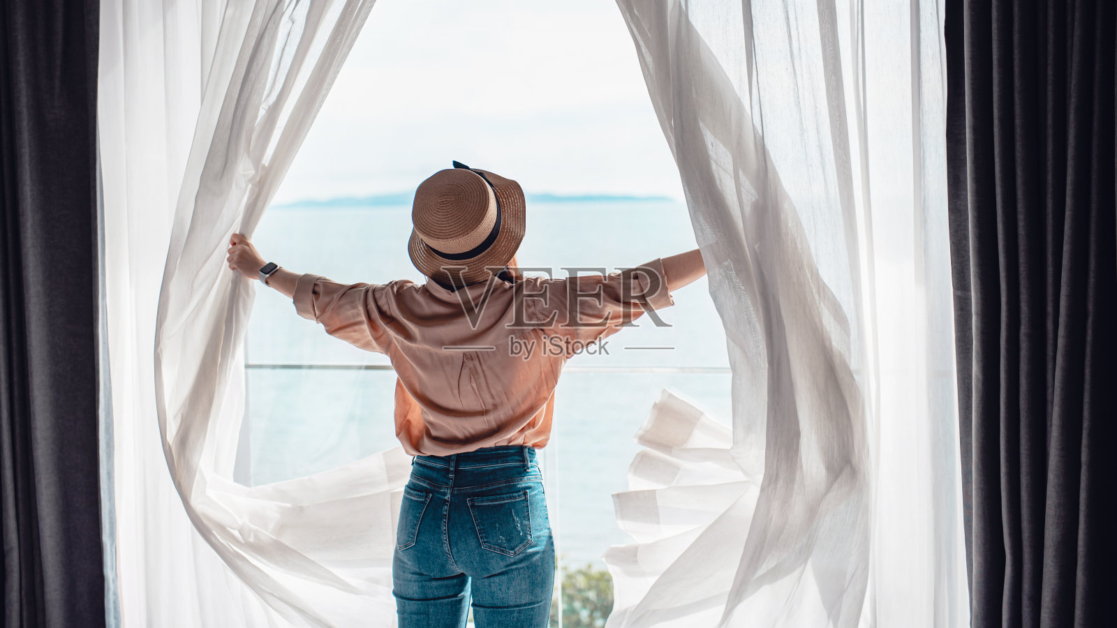 后视图女人打开白色窗帘享受海景，快乐的旅行者呆在高品质的酒店。照片摄影图片