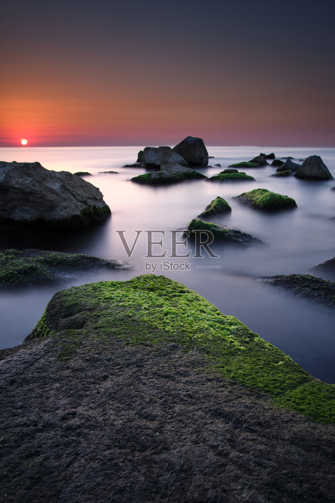 在Badalona海滩上长时间的日出与一些岩石和丝绸效果的水。照片摄影图片