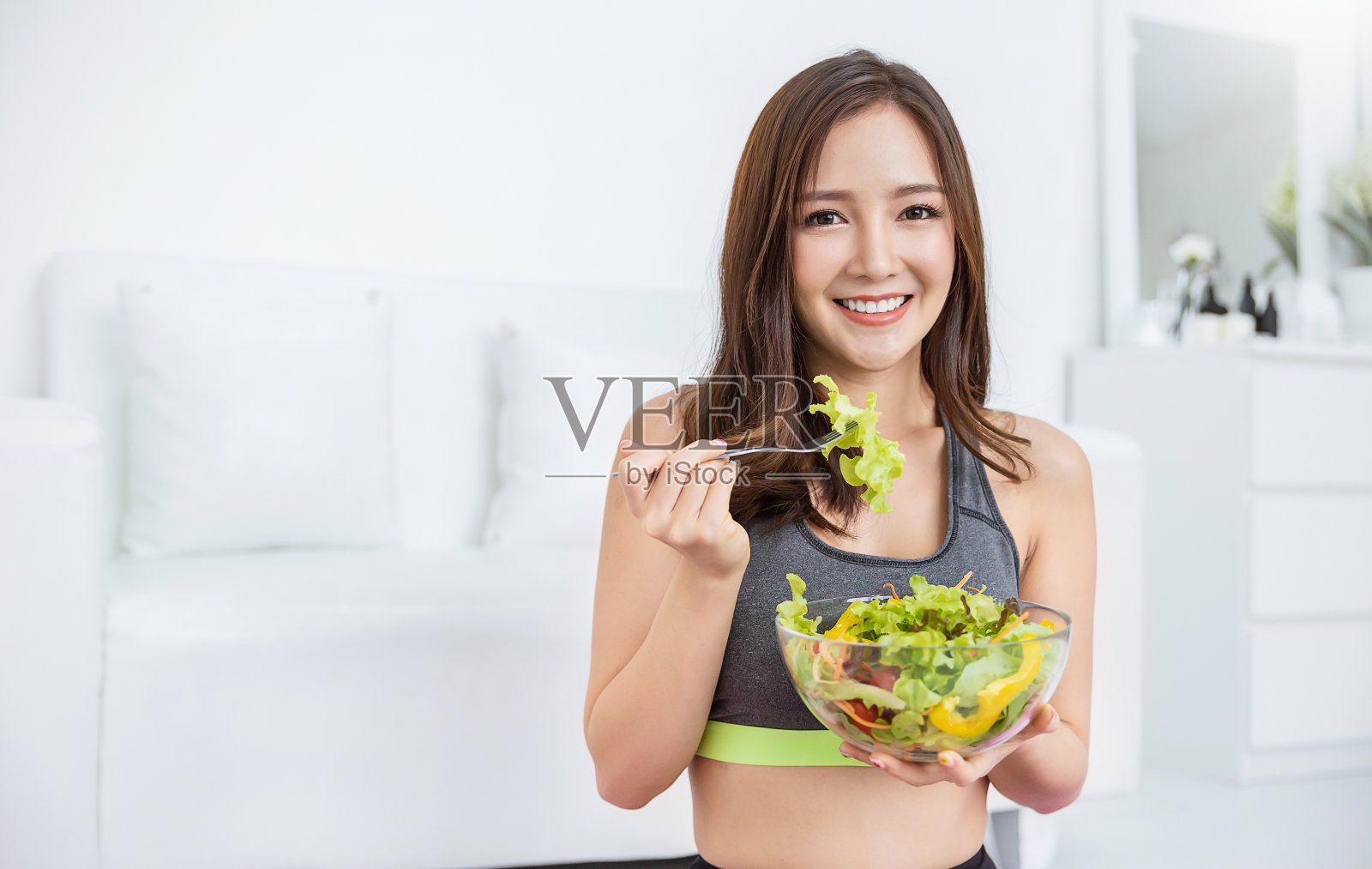 年轻美丽的亚洲女孩的肖像穿着运动服拿着一碗沙拉叉在她的手。素食理念女性吃健康食品和美味食品保健药用植物基地理念。照片摄影图片