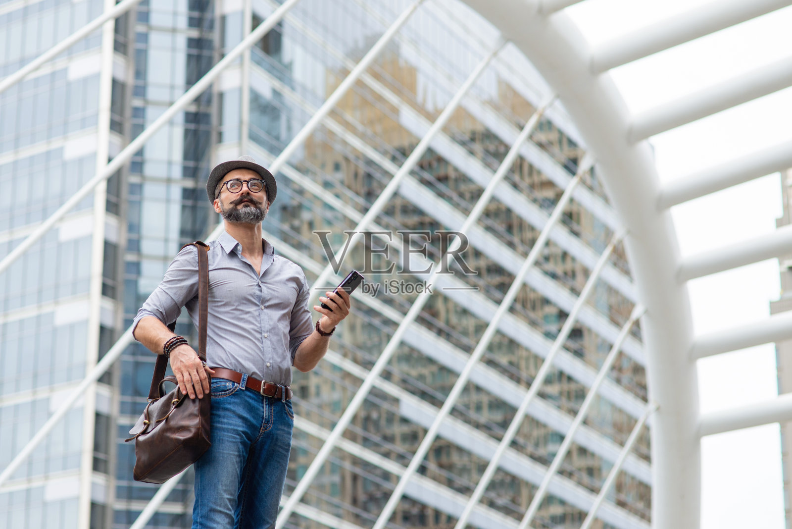 英俊的白人游客在城市中行走使用智能手机与互联网导航地图或在线信息。大胡子的家伙背着皮包，用手机上的应用程序在市区叫出租车。照片摄影图片