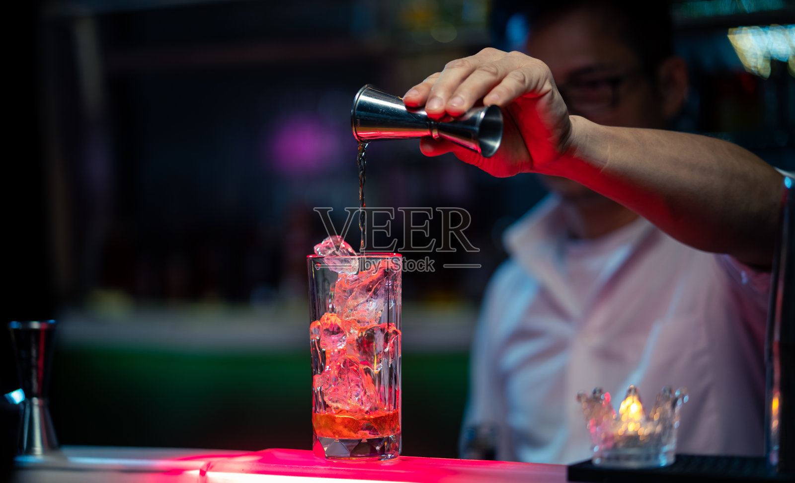 职业亚洲酒保，在吧台将酒倒入加冰块的长玻璃杯中，为夜总会服务。男调酒师调酒师准备酒精鸡尾酒饮料与烈酒架子的背景照片摄影图片