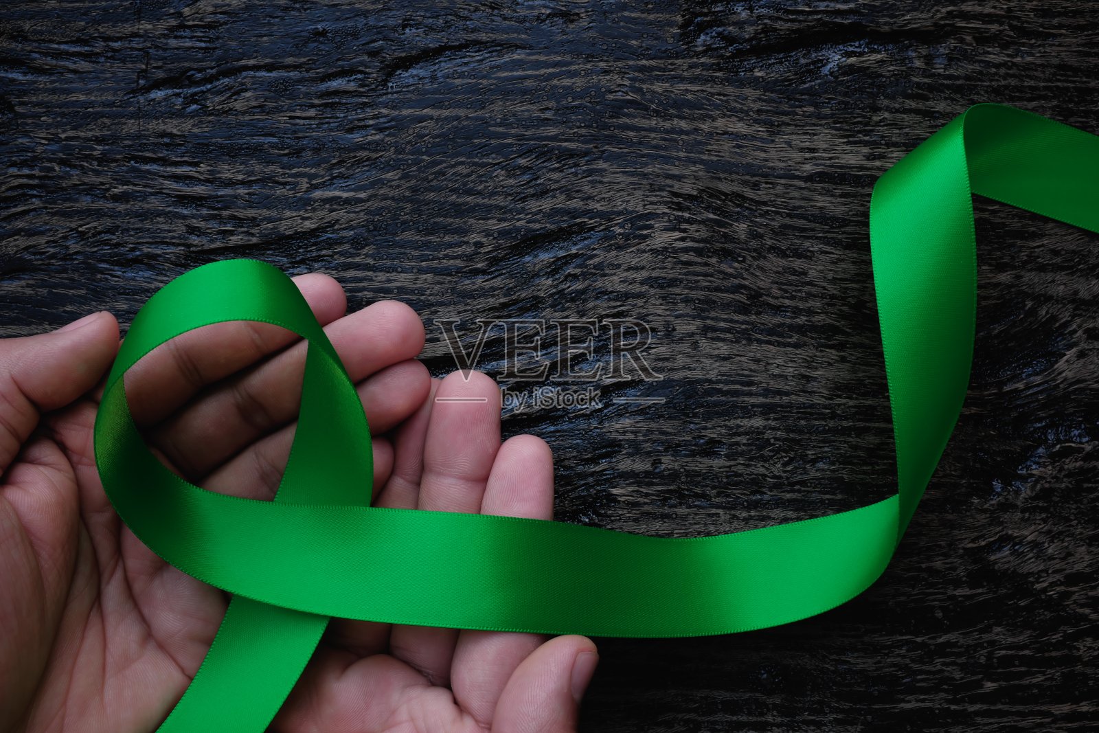 俯视图的手握绿色丝带在黑暗的背景。胆囊癌、心理健康、脑瘫意识、器官捐赠、世界肾脏日和环境保护。照片摄影图片