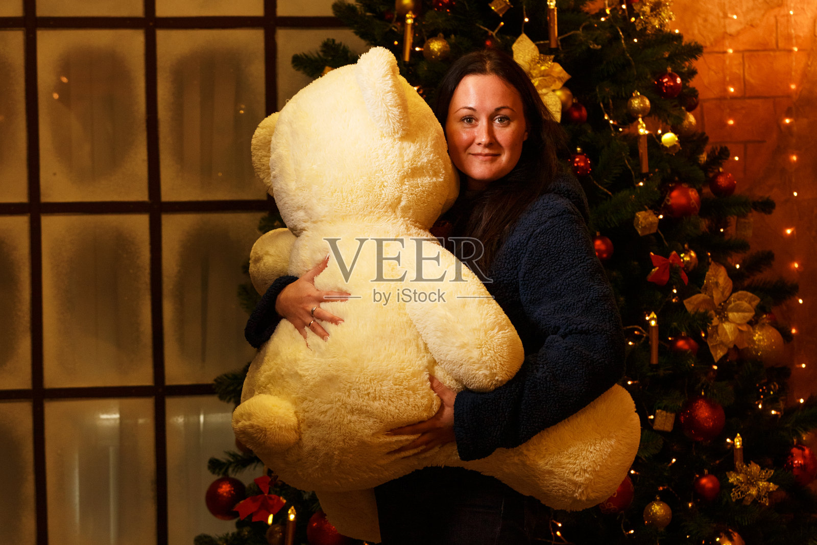 在圣诞树的背景下，一个快乐的女人抱着一个大填充熊玩具照片摄影图片