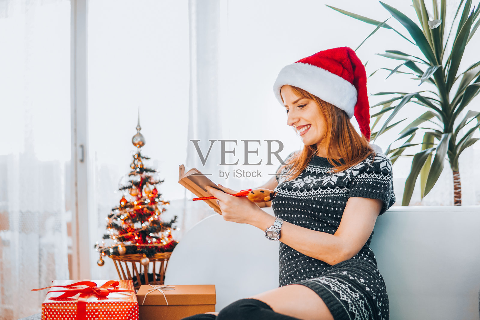 快乐的女人拿着圣诞老人的红帽和圣诞礼物读着圣诞书，打开礼物照片摄影图片
