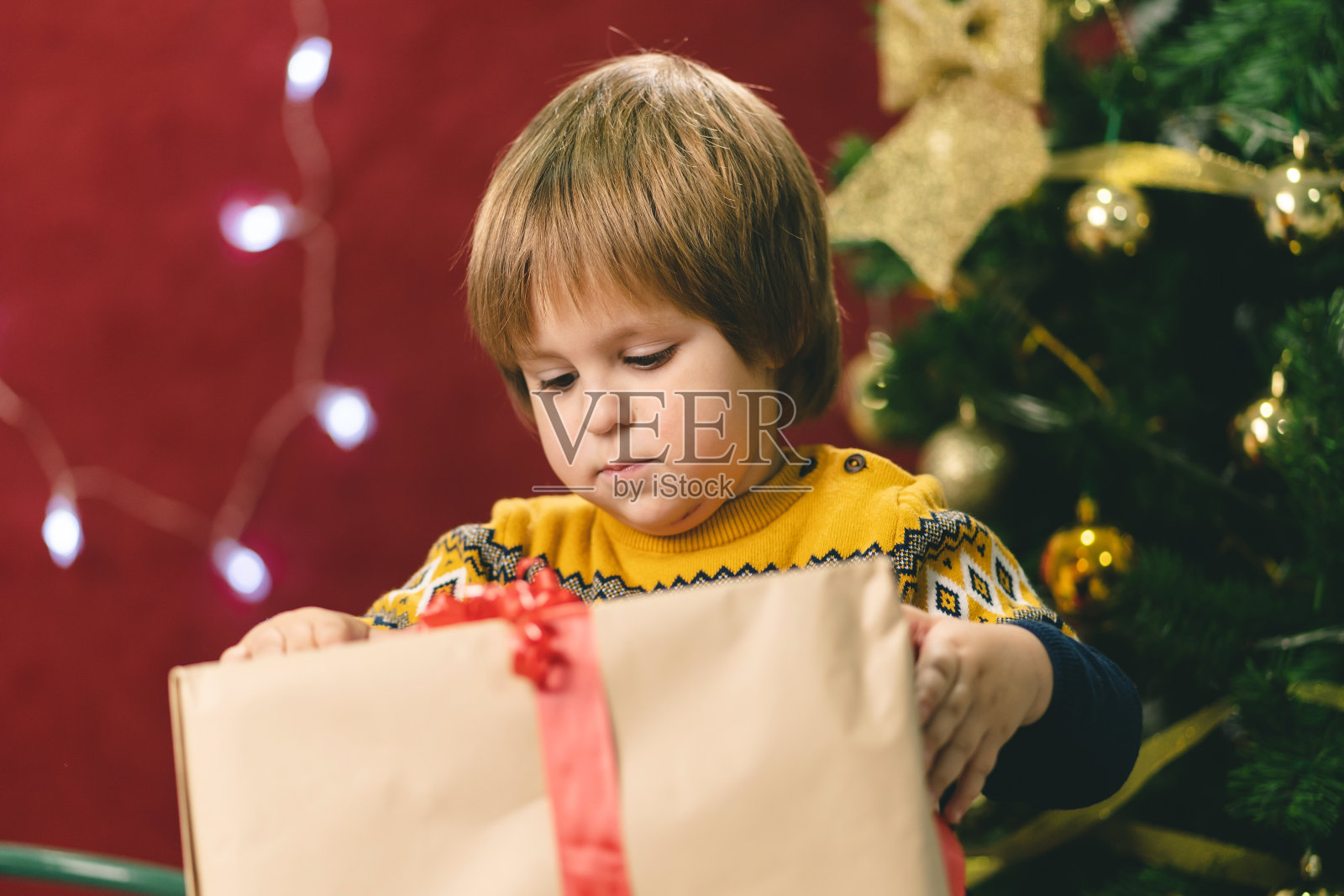 小男孩坐在圣诞树旁打开礼物盒照片摄影图片