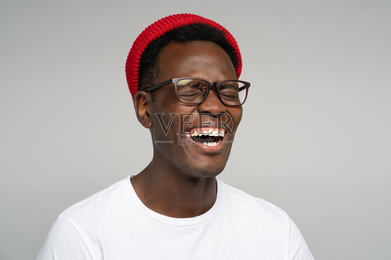 大笑积极的年轻美国黑人男子戴着红帽子心情好，闭着眼睛在镜头前摆姿势。戴着眼镜的黑人男子喜出望外，露出了她灿烂的笑容，孤独的，工作室灰色的背景。照片摄影图片