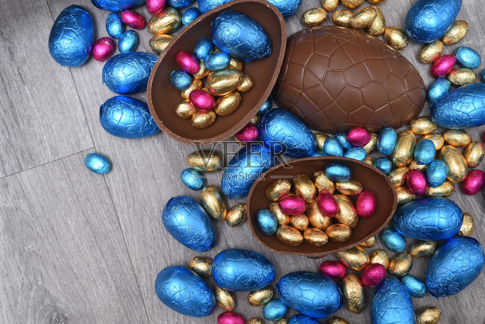 一堆或一组多颜色和不同大小的彩色锡箔包装的巧克力复活节彩蛋在粉红色，蓝色和金色。一个大的半块棕色牛奶巧克力鸡蛋，里面有迷你的鸡蛋，在灰色的木头背景上。照片摄影图片