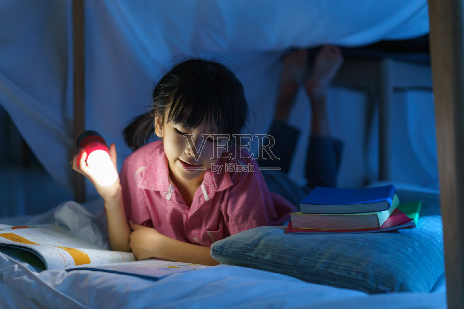 亚洲女孩做了一个营地，在家里的客厅里用手电筒玩想象和看书。照片摄影图片