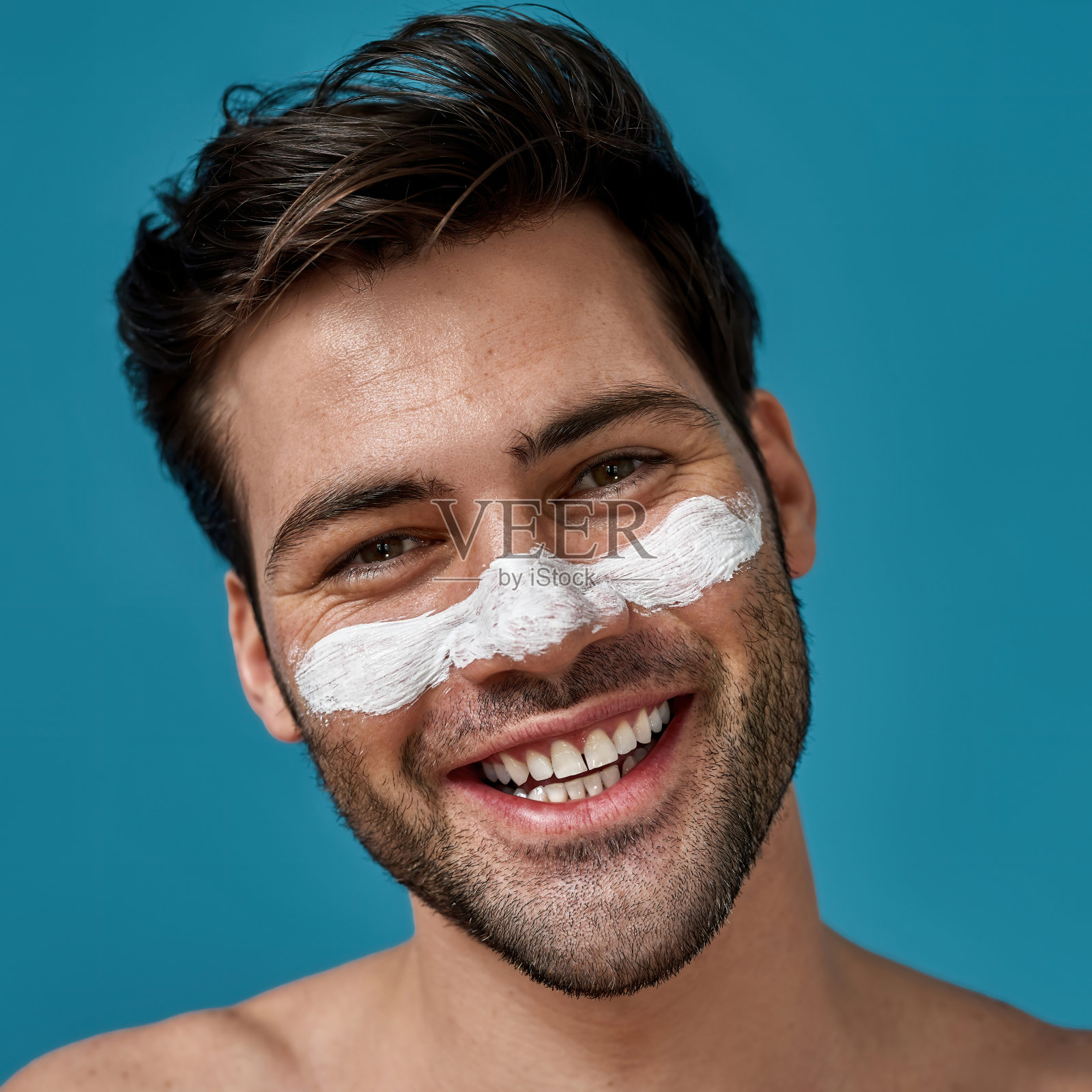 快乐的长胡子的家伙，干净的皮肤和白色的面膜敷在鼻子上，清洁毛孔，微笑在摄像头隔离在蓝色的背景照片摄影图片