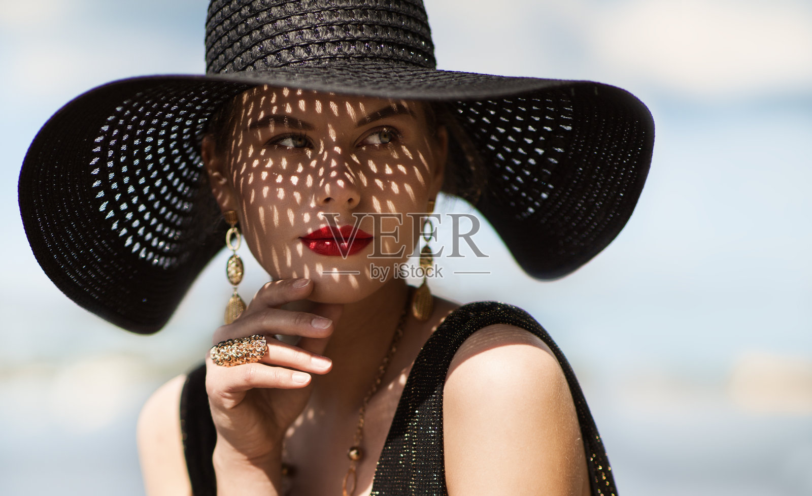 女人在帽子肖像。时尚奢侈模型在黑色夏天帽子与化妆和黄金珠宝。近距离美丽的脸在天空背景照片摄影图片