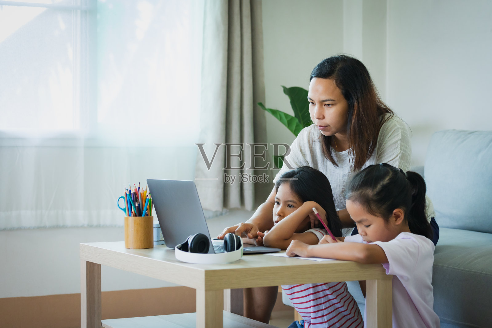 亚洲母亲支持和帮助她的两个女儿在客厅的笔记本电脑上学习。由于Covid - 19大流行，兄弟姐妹在隔离期间在家上学。照片摄影图片