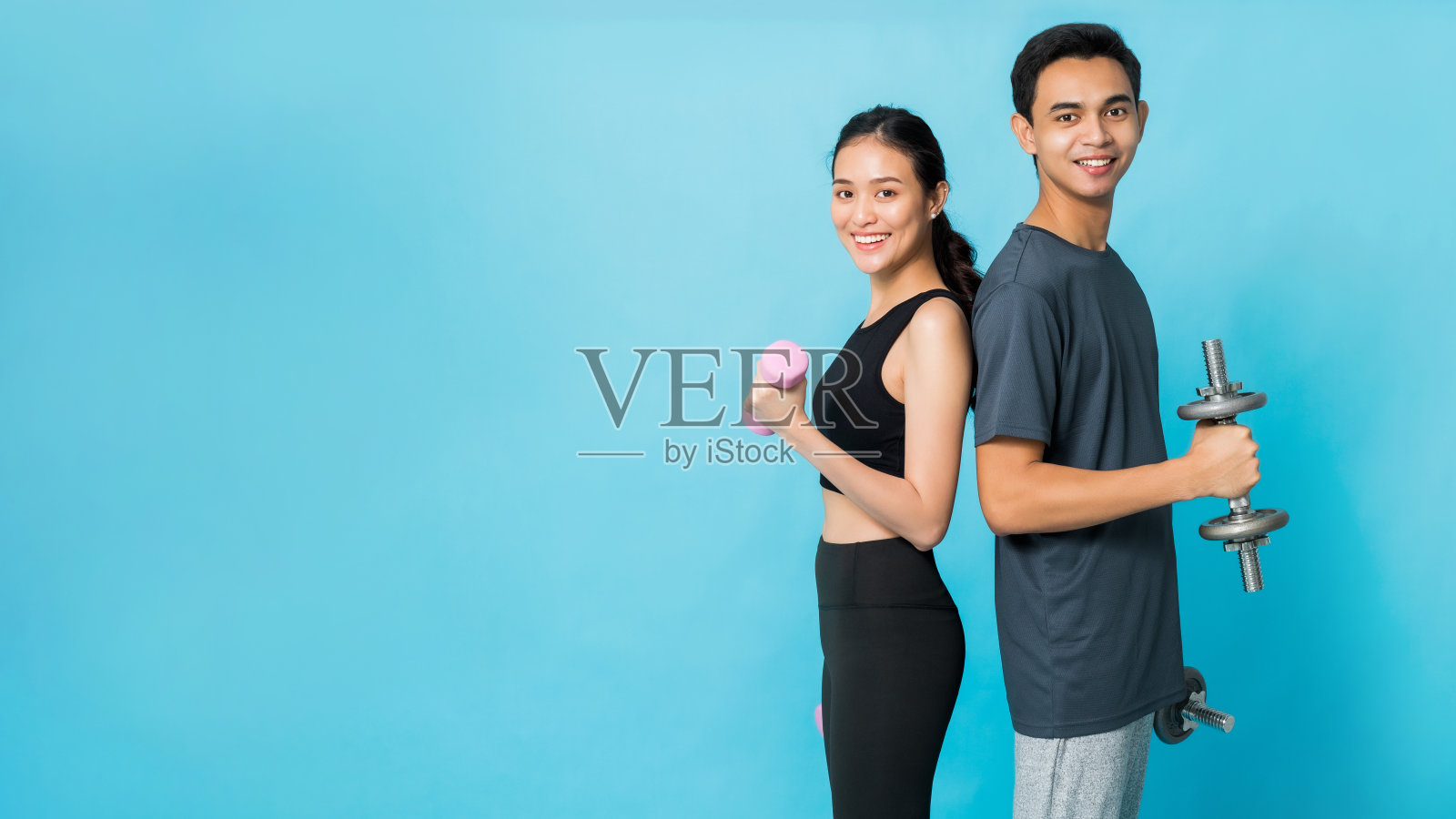 亚洲美丽快乐的女人拿着哑铃，和男朋友站在一起，在蓝色的背景上孤立地锻炼。概念苗条和健康的女孩锻炼。照片摄影图片