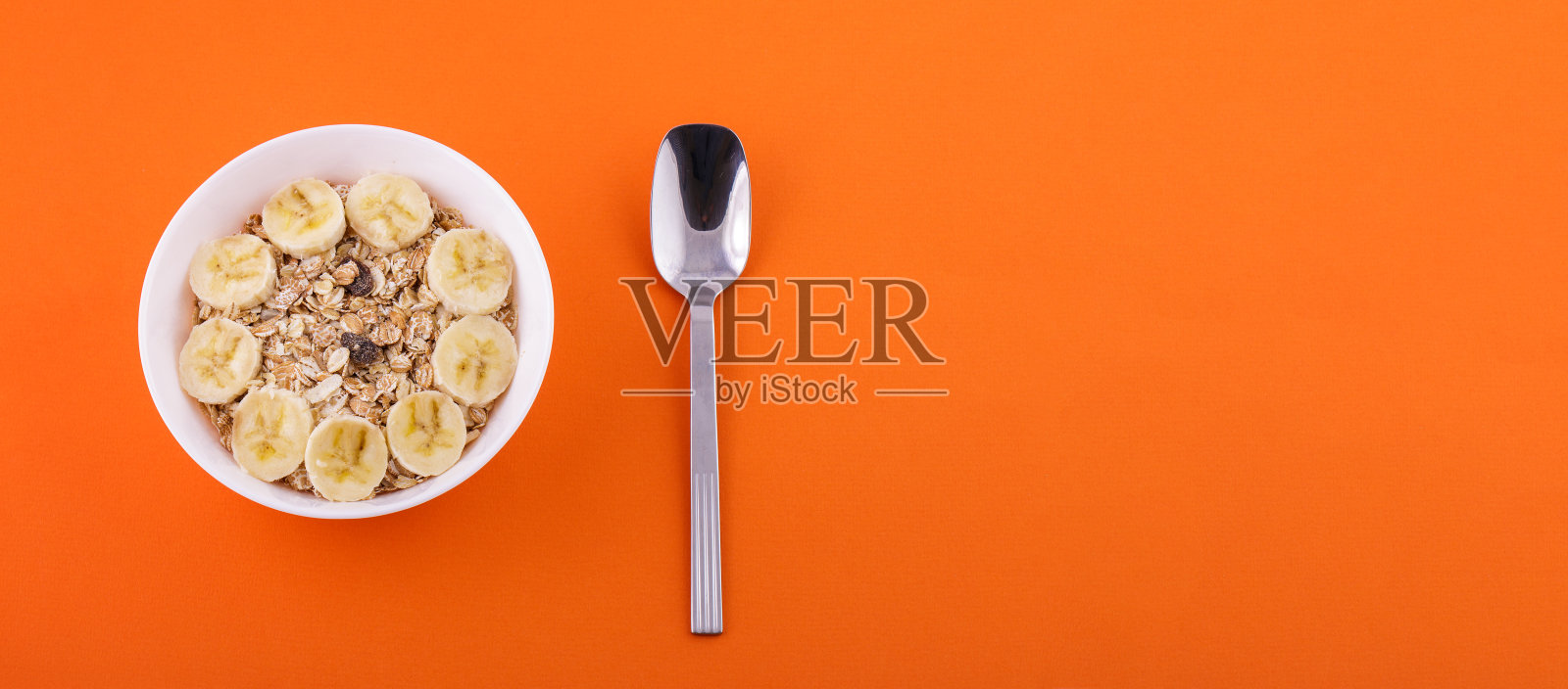 白盘子里的燕麦片配香蕉片和勺子，橙色背景，健康的食品理念照片摄影图片