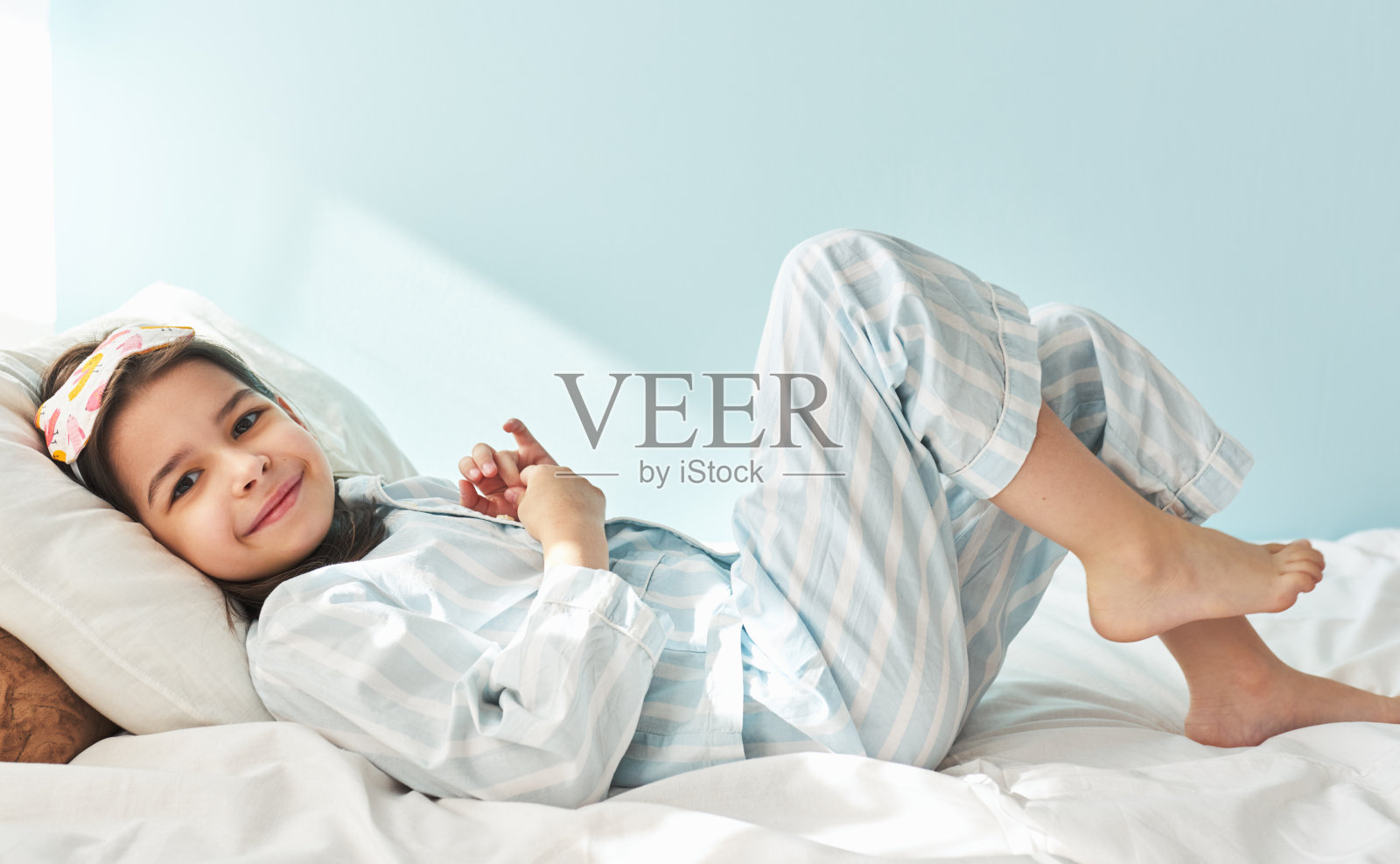 可爱的小女孩穿着蓝白条纹的睡衣，微笑着，看着镜头，躺在床上休息。孩子在早上有快乐的表情。照片摄影图片