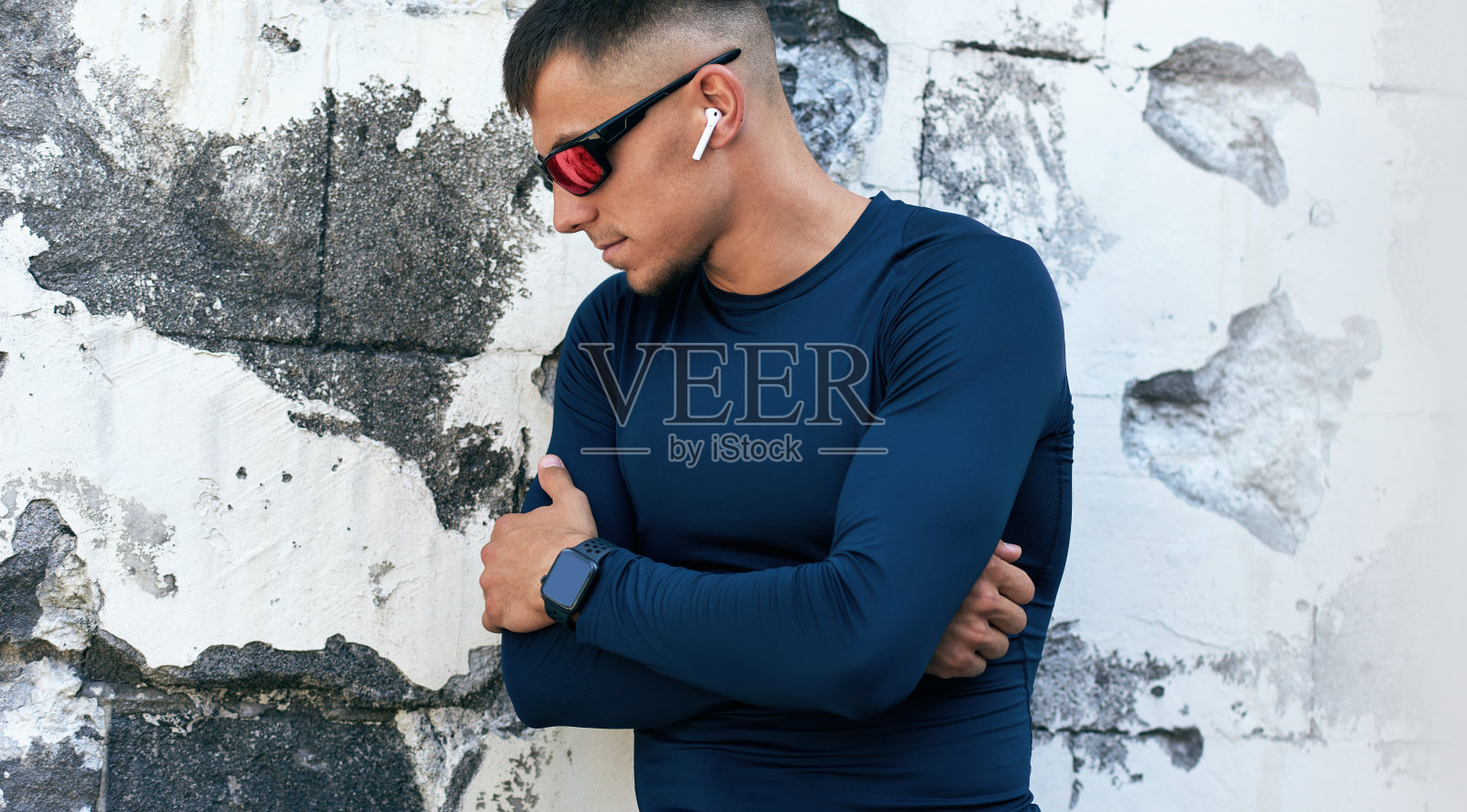 肌肉发达的年轻人，穿着蓝色运动服，戴着红色太阳镜，看着别处，站在城市的混凝土墙。白种运动员交叉双臂休息。照片摄影图片