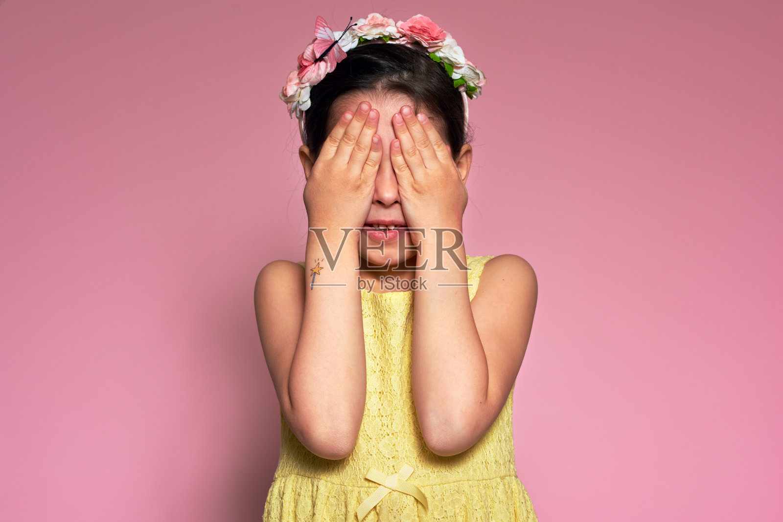 一个小女孩的肖像用双手遮住她的眼睛，在粉红色的背景上摆姿势。可爱的孩子在玩躲猫猫。照片摄影图片