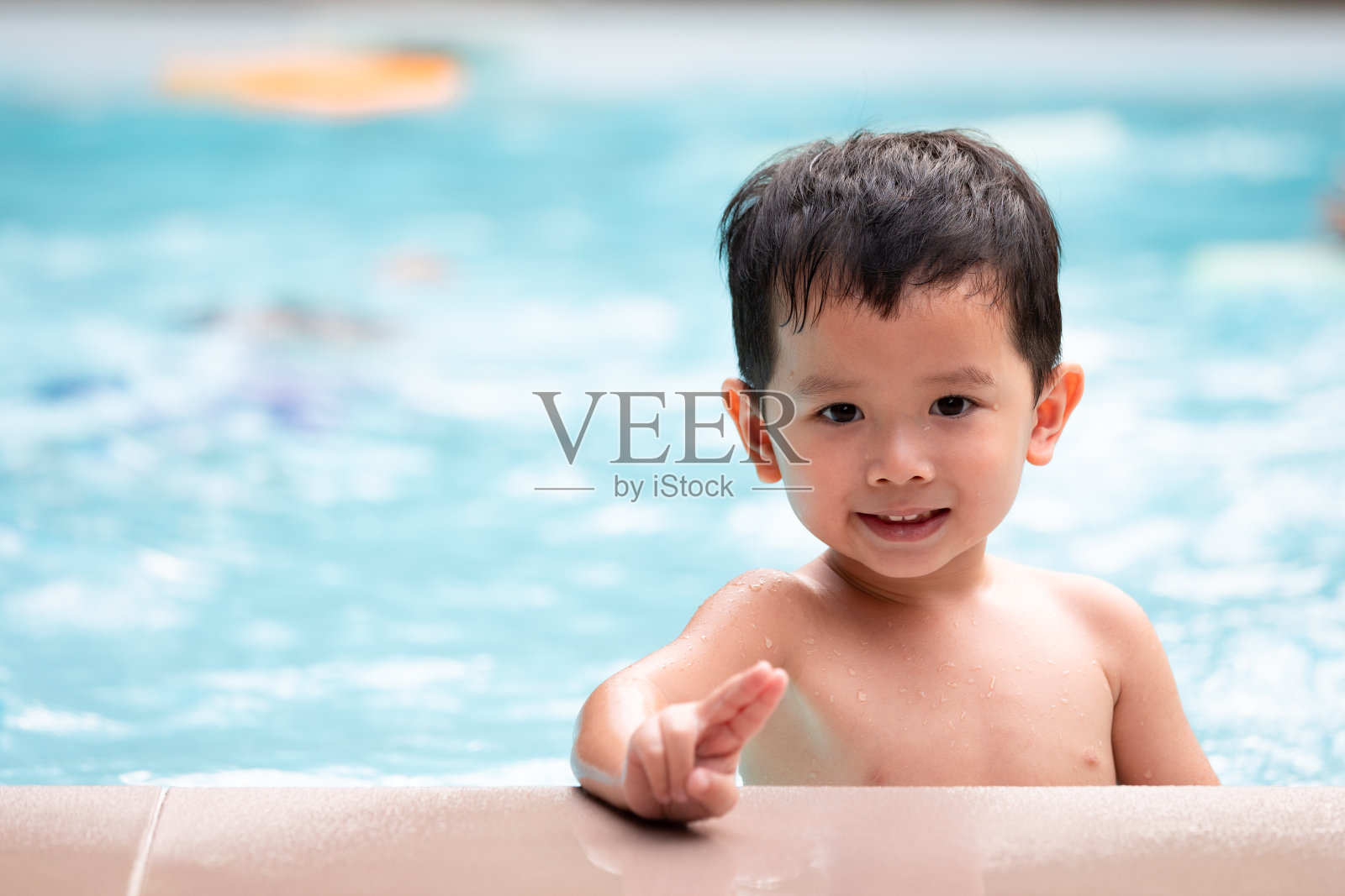 亚洲小男孩的肖像看着相机和微笑，而玩水在游泳池。照片摄影图片