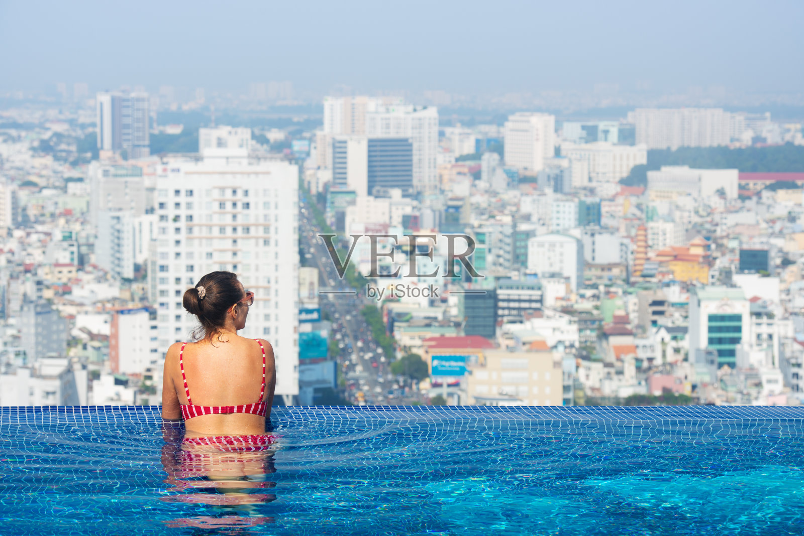 在大城市背景的酒店屋顶游泳池享受年轻女子的后视图照片摄影图片