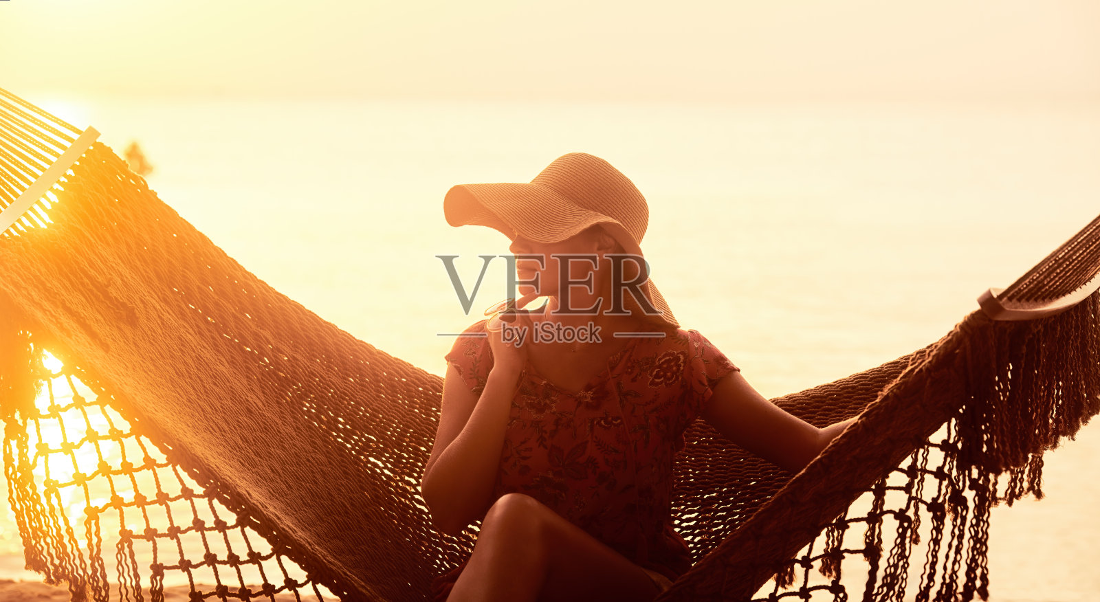 度假海滩夏季度假概念。日落时分，戴着帽子的年轻漂亮女人坐在吊床上照片摄影图片