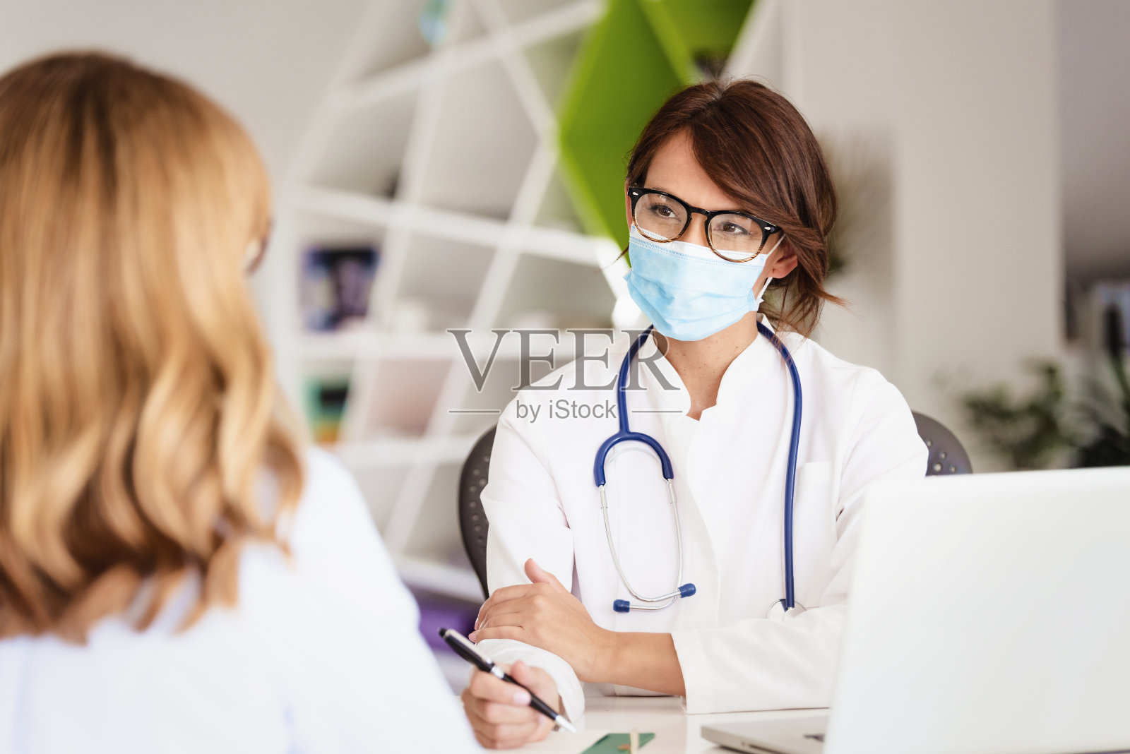 女医生戴着口罩，坐在医生办公室的办公桌前咨询病人照片摄影图片