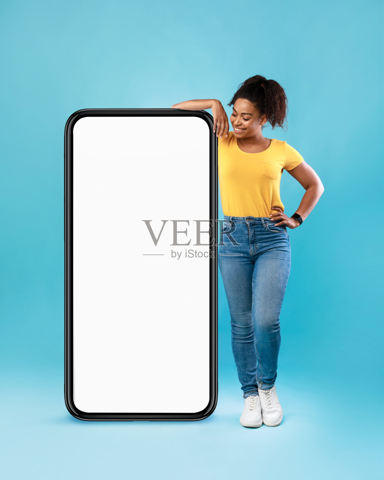 快乐的非洲女人在休闲穿着倚靠在巨大的手机与空白的白色屏幕上的蓝色背景，模型照片摄影图片