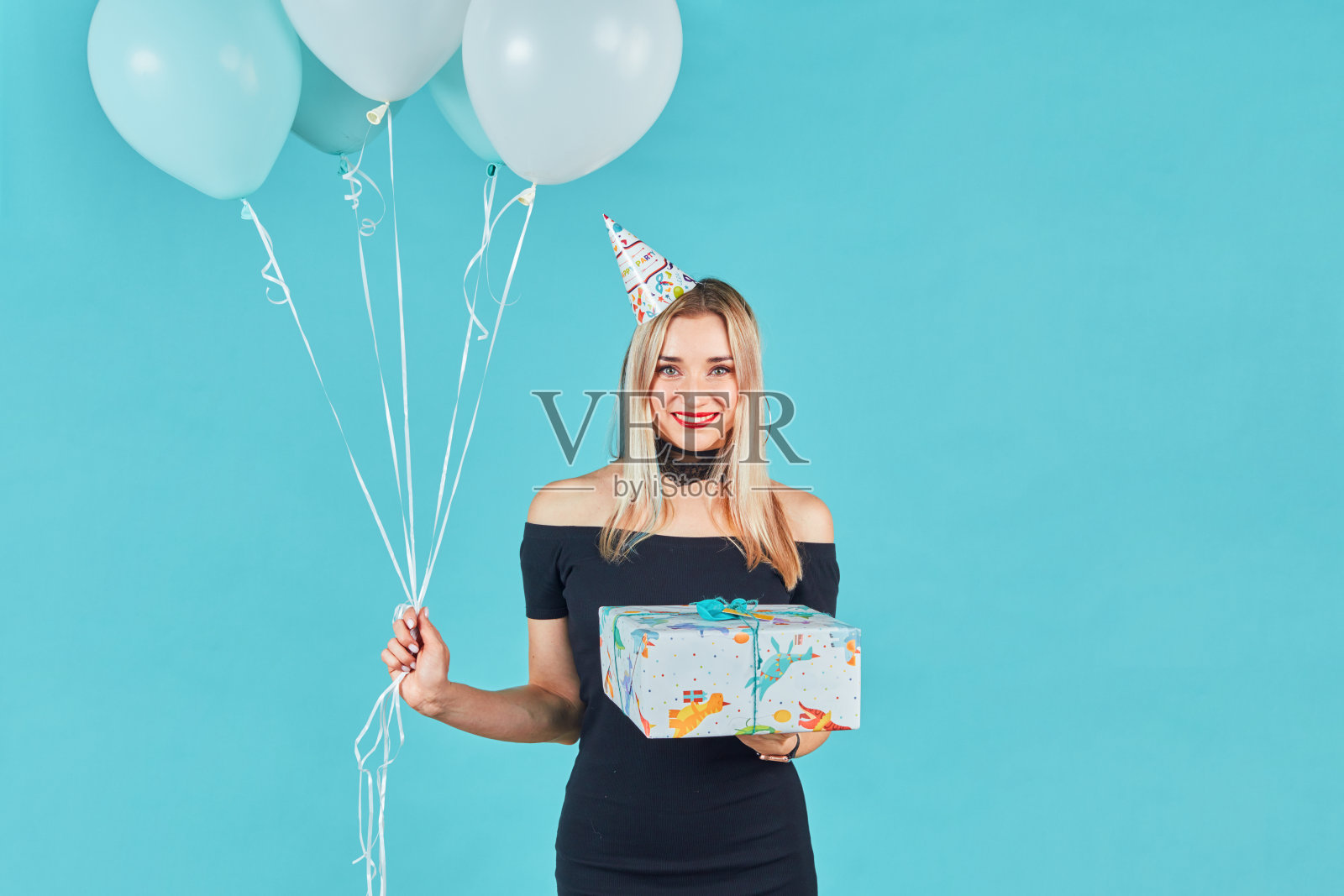 美丽微笑的女人身穿黑色连衣裙，头戴生日帽，手里拿着礼品盒和蓝色背景下的彩色气球。可爱快乐的年轻女孩庆祝生日派对。照片摄影图片