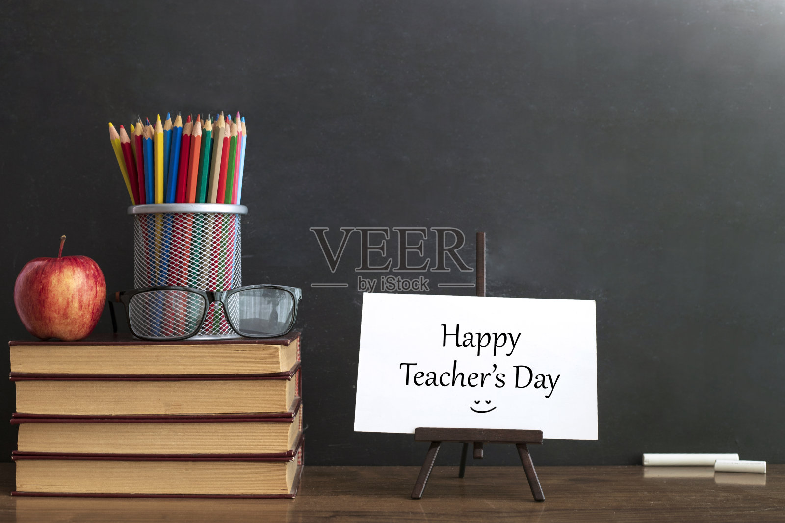 卡片上写着:教师节快乐。眼镜老师的书和一个站在桌子上的铅笔，在黑板的背景上用粉笔。教师节的概念。照片摄影图片