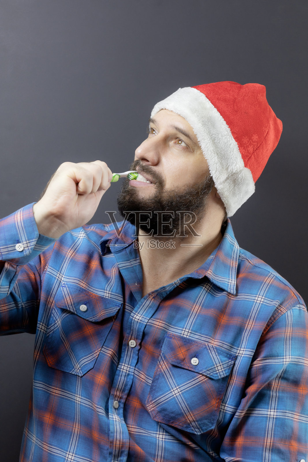 一个戴着一顶奇异帽子、满脸胡须的男人正在用绿色牙刷刷牙。为圣诞节做准备。黑色的长胡子的圣诞老人照片摄影图片