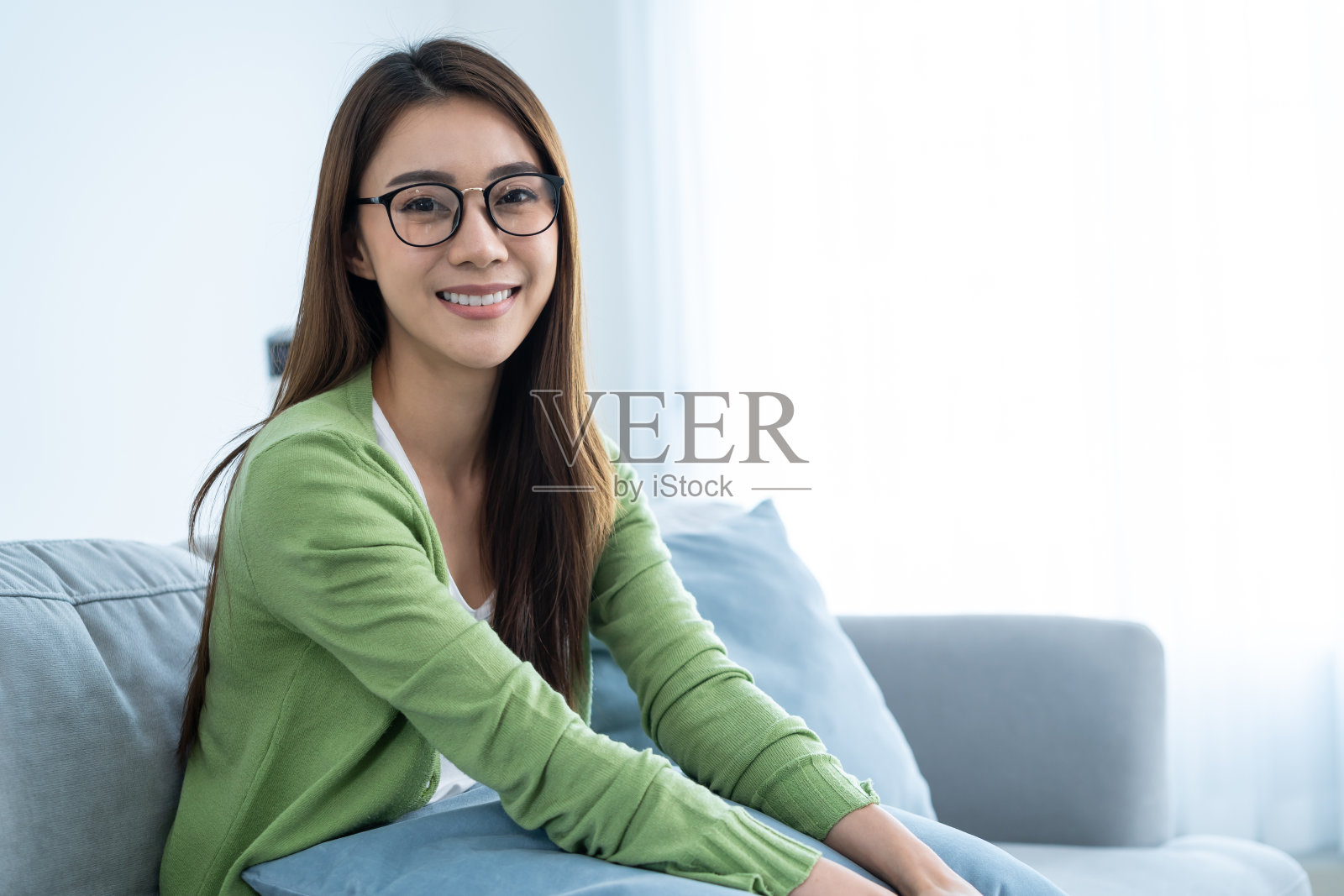 亚洲美女的肖像坐在家里客厅的沙发上。漂亮的年轻女性戴上眼镜，心情愉快，放松，享受清晨周末在家活动的生活方式和微笑。照片摄影图片