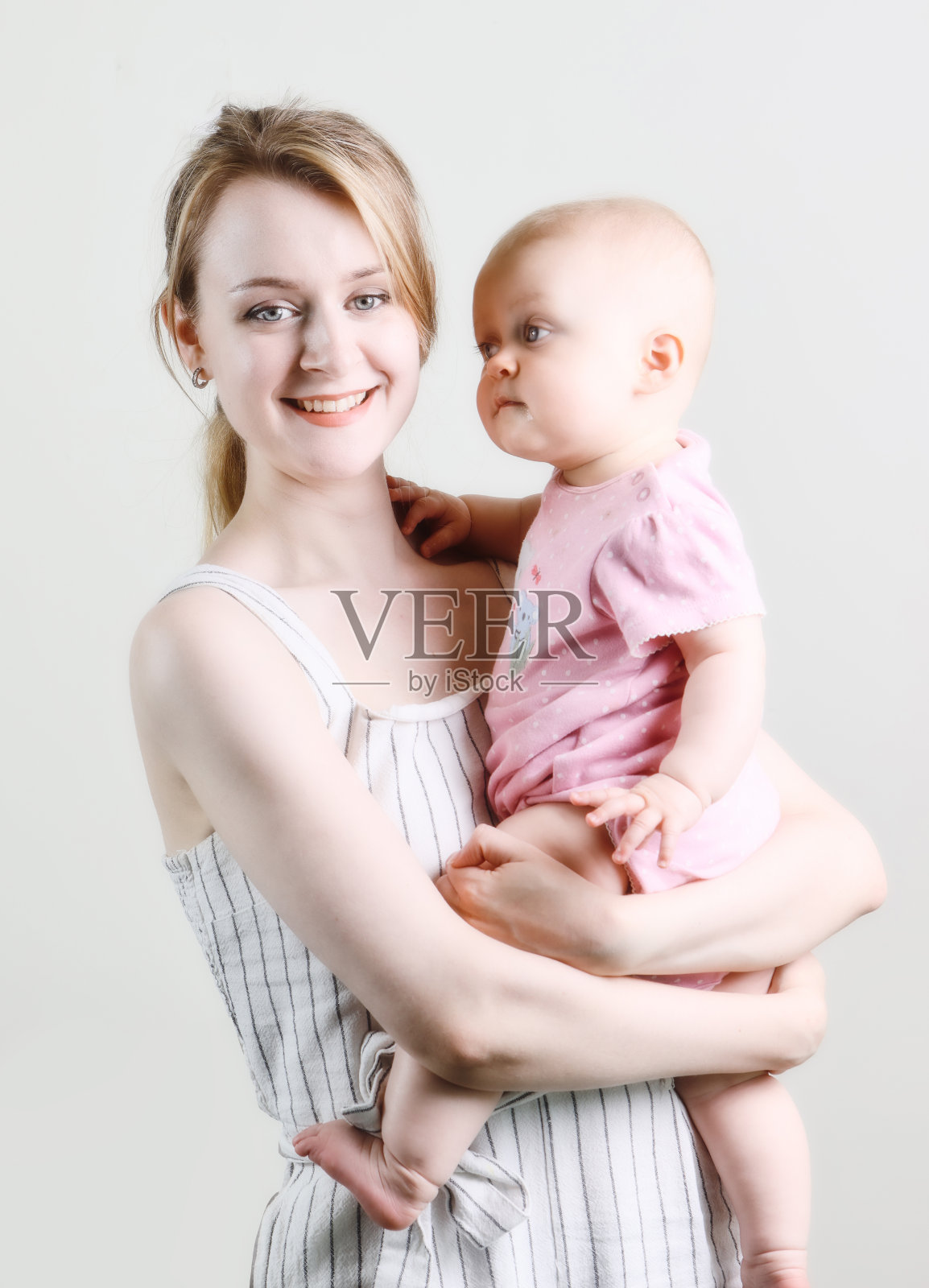 妈妈对着镜头微笑，她抱着9个月大的女婴，女婴好奇地看着她。拍摄在朴素的灰色背景照片摄影图片