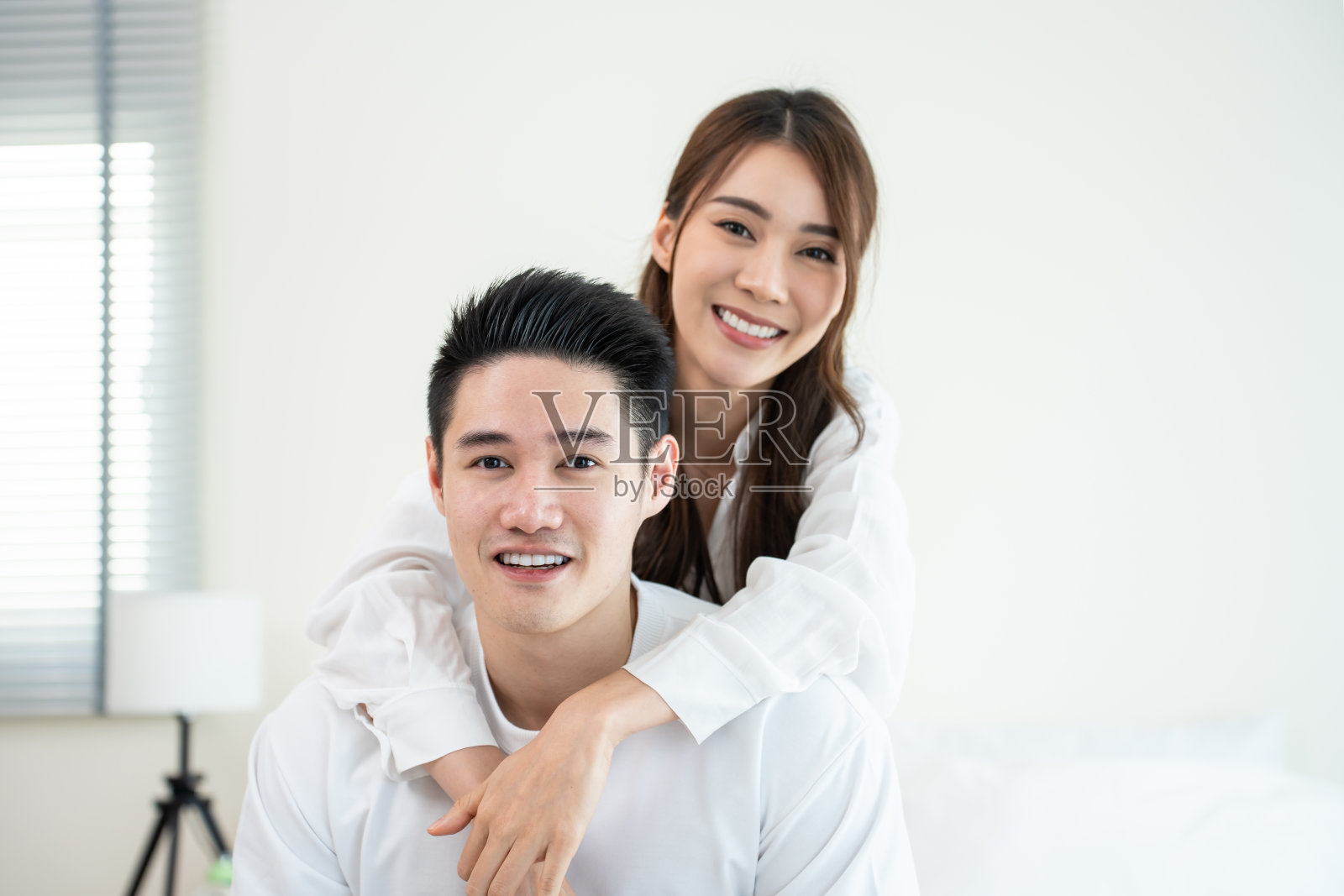 亚洲新婚夫妇的肖像坐在床上，看着相机。漂亮漂亮的年轻男女穿着睡衣在家里的卧室里享受清晨的活动。家庭关系的概念。照片摄影图片